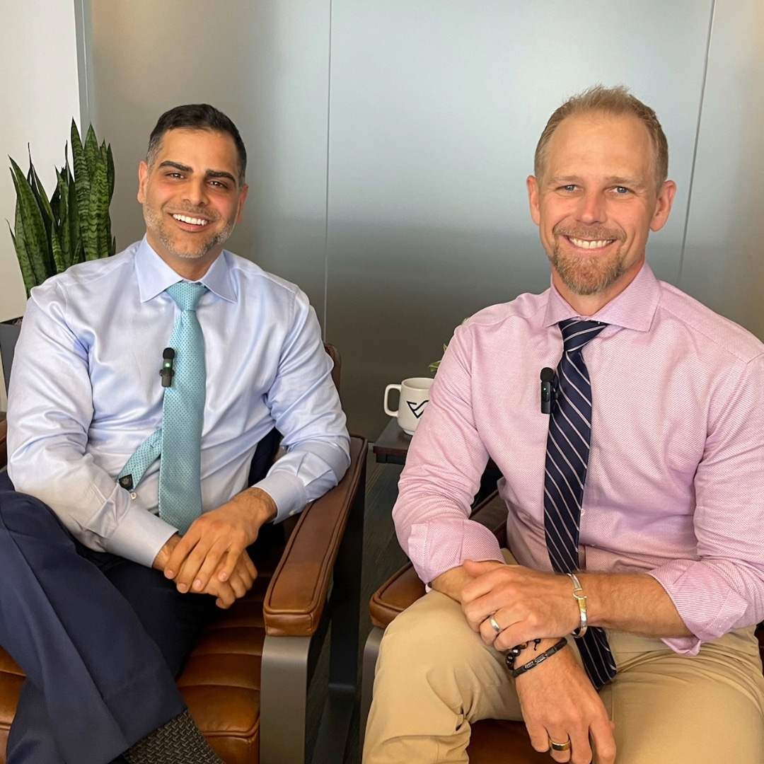 Man in blue shirt and man in pink shirt sitting for a podcast