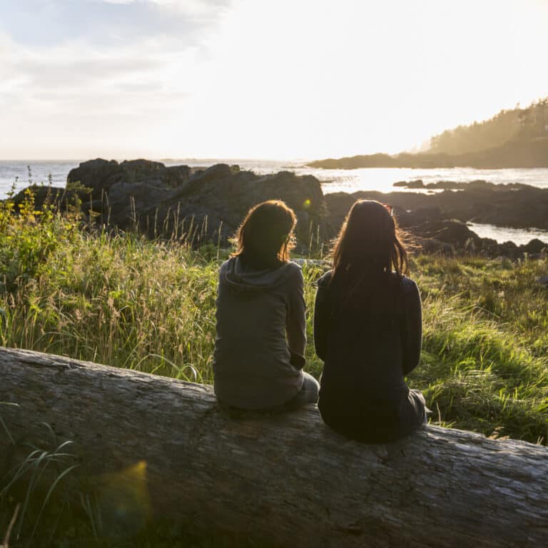 two women relax on log, look out across bay