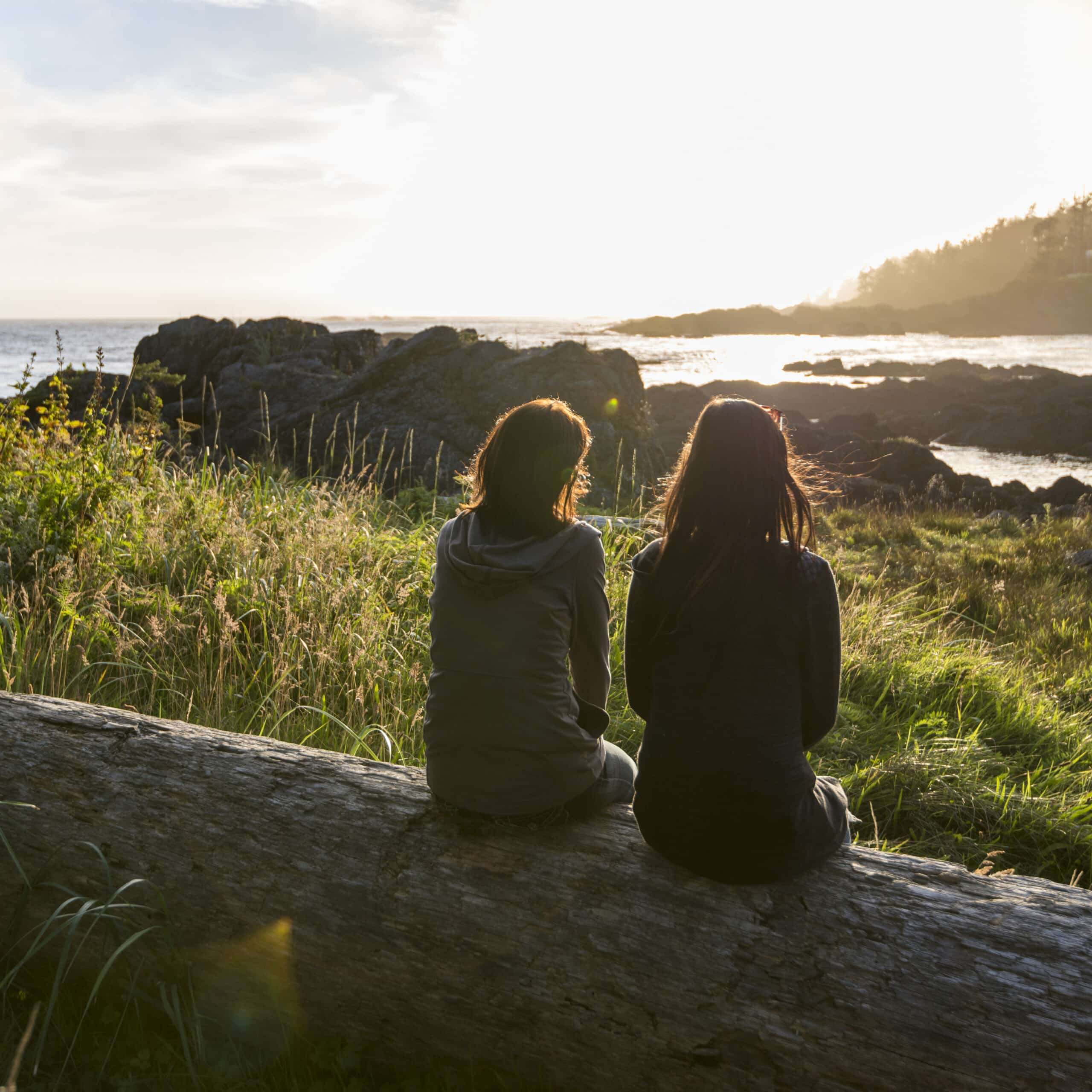 two women relax on log, look out across bay